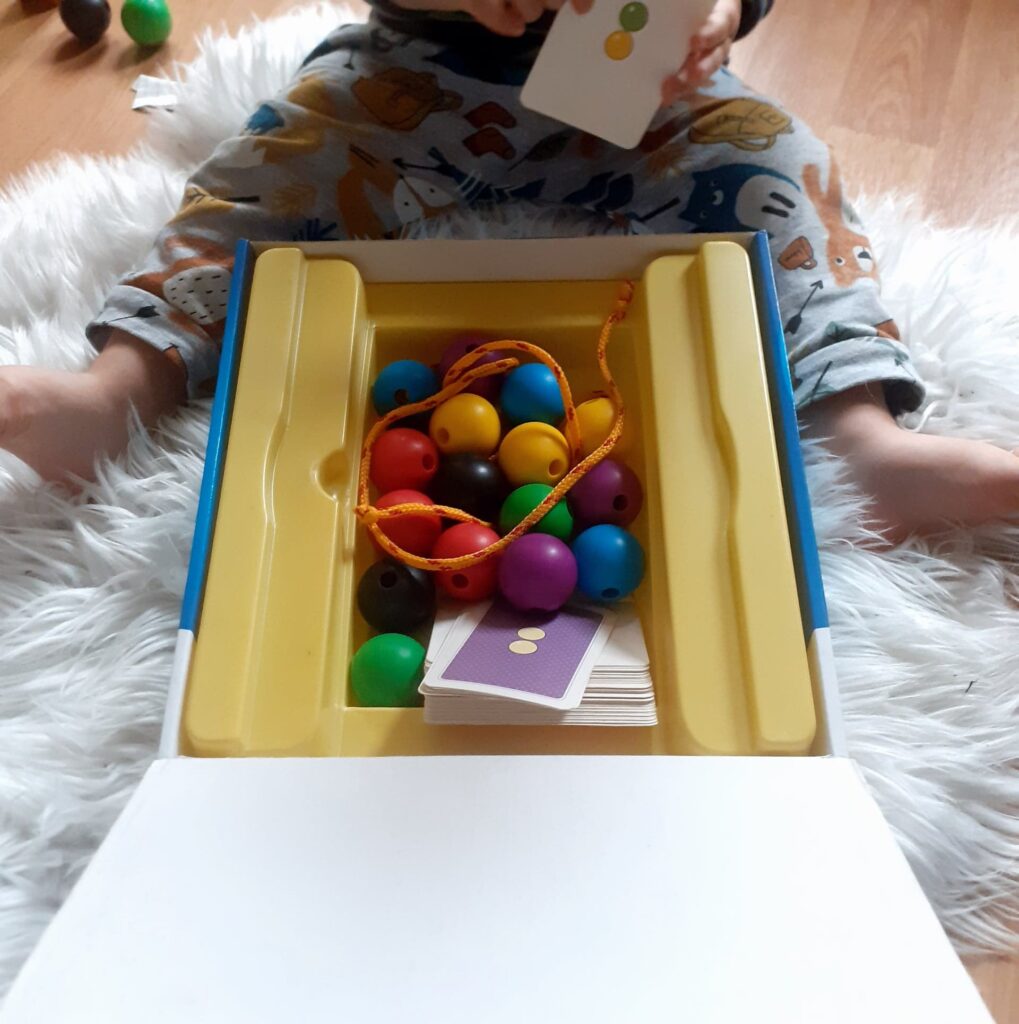 Dziecko bawiące się zabawką "Nawlekaj nie czekaj". W pudełku kilka kolorowych kulek do nawlekania wraz ze sznurkiem oraz etykietami.